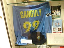 A blue coloured T-shirt displayed at a store window. The T-shirt has the words "Ganguly" and the number 99 below it, both in yellow colour. Beside the T-shirt, a picture and an open book is visible.