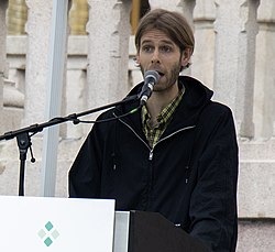 Lars Vaular, en mann med brunt hår og skjegg, står og snakker inn i en mikrofon.