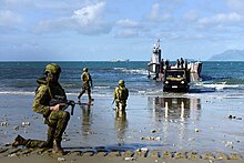 Colour photograph of three men wearing green military uniforms kneeling on a beach near a grey ship. A green truck is driving off the ship, and a large grey ship is visible on the horizon.