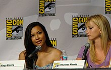 A dark-haired woman is watched by a blonde woman as she speaks at a convention panel.