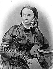 Photographie noir et blanc d'une femme blanche en buste, cheveux sombres en bandeaux, assise, une main posée sur la joue, l'autre tenant un livre, toilette sombre