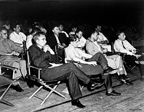 A group of formally dressed people sit in the audience, on folding chairs, and listen to a lecture