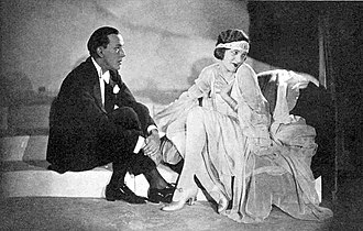 young white man and young white woman in evening costume, seated; he is looking intently at her; she is looking away coyly