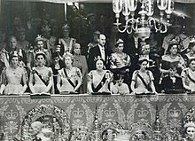 Three rows numerous men and women in fancy dress. Many of the women are wearing tiaras and sashes with white dresses