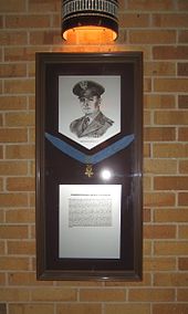 A plaque in a glass enclosed frame depicts a black and white image of a soldier in WWII regalia, a medal of honor suspended across the middle, and a description of the event for which the individual was awarded the medal
