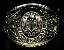 Close-up of gold ring. On the top, the words Texas A&M University 1876 encircle an eagle atop a shield over the numbers 0 and 4. The left side contains a large star and an oak. The right shows a cannon, saber, and rifle with the crossed flags of the United States and Texas.