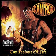 An image of Cam'ron wearing overalls but no shirt. He has googles on his head and is wearing heavy worker gloves to hold a sledgehammer. He appears to be in a smeltery or foundry and in the background a vat is pouring molten metal, causing sparks to fly.