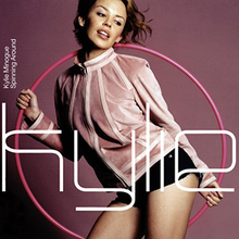A woman dressed in a rose-coloured jacket and black hotpants is striking a pose in front of a dark purple background with a pink hoop in her hand. The word "Kylie" is written in front of her in a large white font.