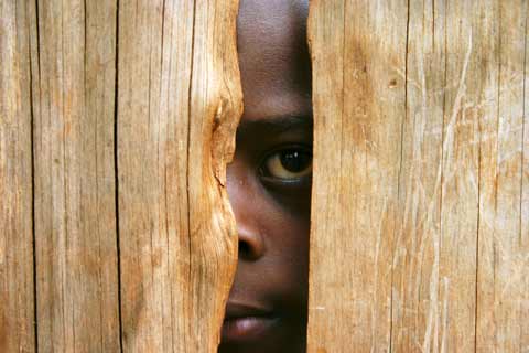 Image of young black boy looking through a gap in a wooden fence