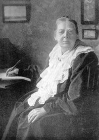 Photographic monochrome portrait of a lady wearing a dark dress with frilly white bib. She is holding a pen, which rests on a pad of paper on a desk by her side.