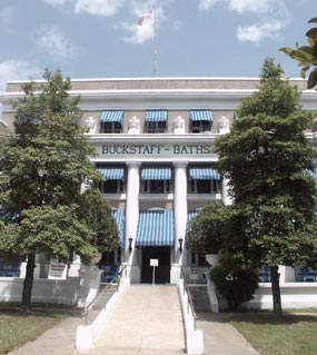 color photo of Buckstaff, showing the ramp and center front of the building. It is a three story building of grayish-brown brick and white Doric colums with classic urns flanking the top of the ramp and between the third floor windows. It has blue and white striped awnings.