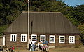 Photograph of the Fort Ross Commander's House on a sunny day. Visitors stand at a table in front of the rectangular log building with a high, peaked roof and white painted windowframes.