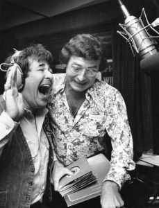 Hy Lit (left) with his hand raised and mouth open, laughing, wearing a denim vest over a white, pocketed, collared shirt with a headset on. Joe Niagara is to his right (center) holding a stack of 45rpm records and wearing large glasses and a jumping-horse-patterned collared shirt. A microphone on a boom arm is on the right.