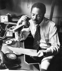 George Woods sits facing left, but toward the camera. He holds a sheet of paper up with the top near the camera, likely reading something on air into the large microphone that sits in front of him on a short stand. A rotary phone is near the front of the frame and a small clock is in the background. Two 45rpm records lay in front of him next to a small two-position switchboard. He has short hair, sideburns and a mustache, and wears a stripes, light-colored or white shirt with a patterned dark tie.