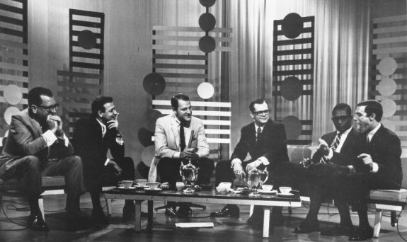 six men sit around a coffee table (with coffee cups and a chromed kettle atop it), all leaning forward in discussion, presumably, about music. the backdrop of the set is decorated with standing wooden slats, like vertical fences and modernist circles. The men all wear suits and ties, most smirking, looking toward Jerry Blavat (far right), who has his arms outstretched and is sticking out his tongue.