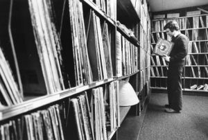 A bookcase-style shelf-lined wall takes up a majority of the frame, coming closer to the viewer. to the right, facing the wall is DJ Jon Takiff, wearing a sweater or jacket over a white collared shirt (with the collar sticking out and over top). He holds and inspects what appears to be a Looney Tunes soundtrack vinyl record sleeve. the rear wall is also lined with shelving and records.