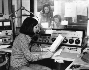 Della Lazarus sits reading a thin booklet facing right, with a microphone in front of her face and controls just behind her. A soundproof window is behind the audio controls and two men sit on the other side, one sitting (likely Bill Sinrich), reading the contents of a paper into a mic of his own.