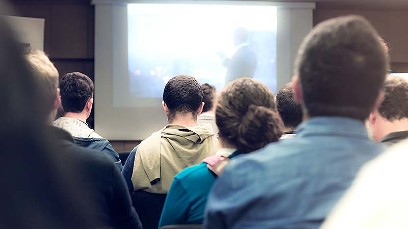 Back view of people in a meeting, looking at a presentation on a screen.