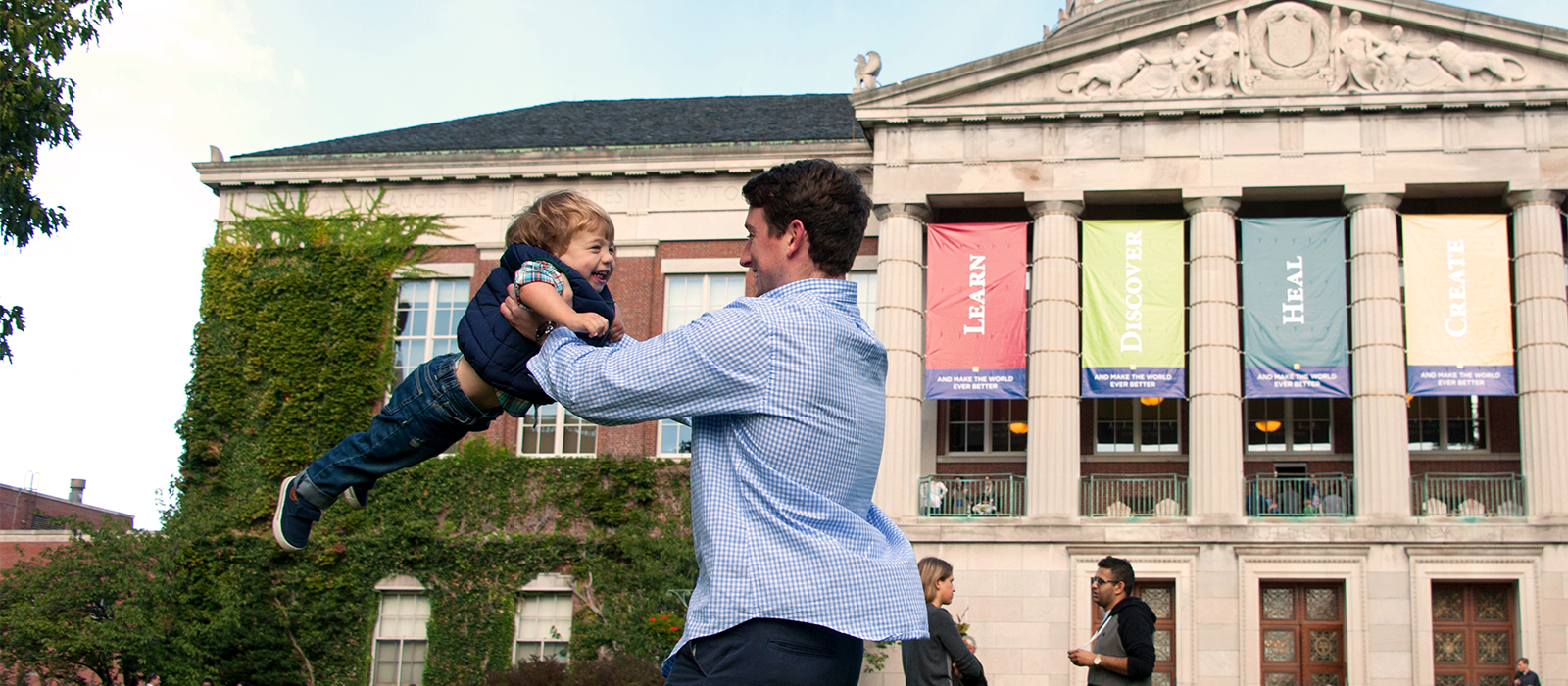 man swinging young son in front of Rush Rhees library