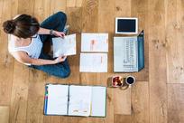 Student sitting on wooden floor surrounded by papers, laptop, digital tablet, file folder, coffee and fruit bowl