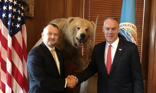 Secretary Zinke shakes hands with Cam Sholly in front of flags in wood panel office.