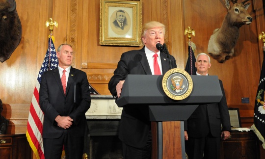 President Trump wears a dark suit and red tie and speaks from a podium in a wood paneled office as Secretary Zinke and Vice President Pence look on.