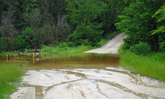A gravel road has thick ruts and is partially flooded where it goes down a hill before going back uphill into a forest.
