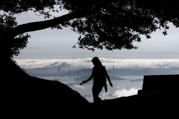 Danielle Roush is silhouetted against a view of fog rolling across the San Francisco skyline and into the bay, as seen from Grizzly Peak Boulevard in Berkeley.