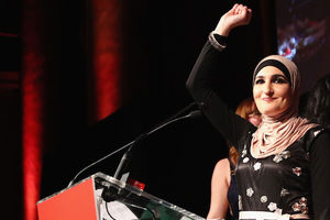 A brown woman wearing a black floral dress and a light peach hijab smiles while holding up a fist