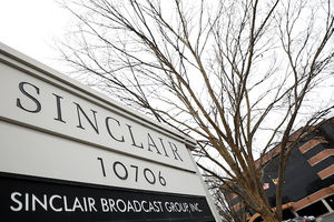 A sign for Sinclair Broadcast Group appears at the forefront of a parking lot, a tree without leaves, and the building for the corporation in the far background with an overcast sky.