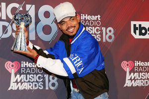 Chance the Rapper smiles in grey hat and blue jacket with white detail while holding silver award in front of red background with white text