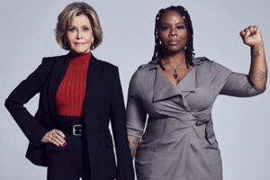 Jane Fonda and Patrisse Khan-Cullors. White woman in navy suit and red sweater holds hands with Black woman in grey dress with black arm tattoos and raised fist, in front of grey background