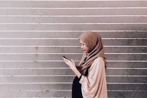 Light brown hijabi woman wears taupe headscarf, tan sweater and black dress, looks down at cell phone in her hands