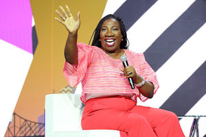 Tarana Burke. Black woman with black dredlocs in pink shirt and pants waves and holds black microphone while sitting on white chair in front of black and white and gold and purple background