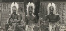 Three Torres Strait men wearing head dresses sitting outside a hut 