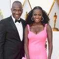 Black man in black and white tuxedo smiles next to Black woman in pink dress in front of white wall with gold ornamentation and insignia