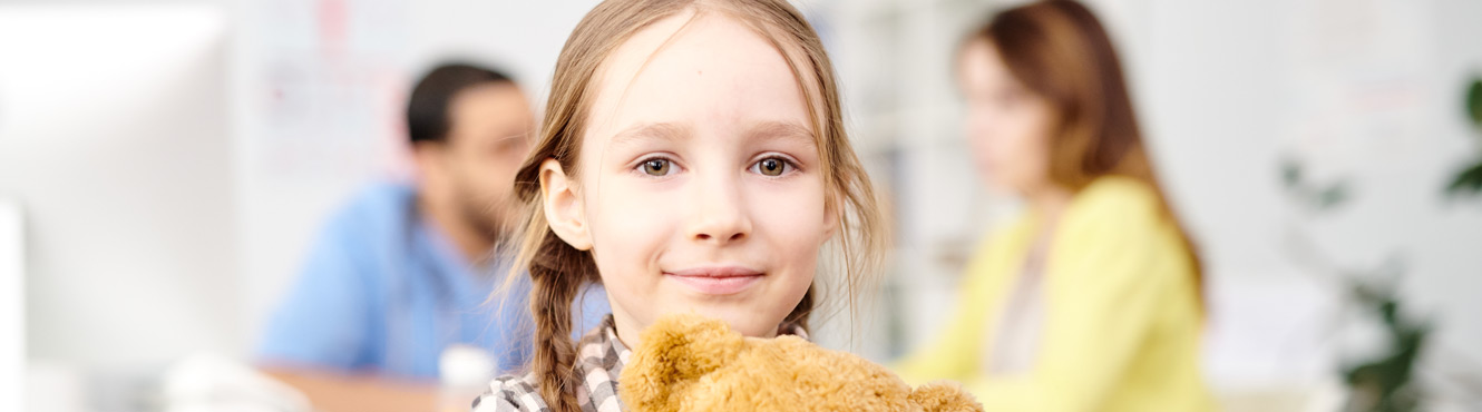 Child holding her teddy bear with parent talking to doctor in background 