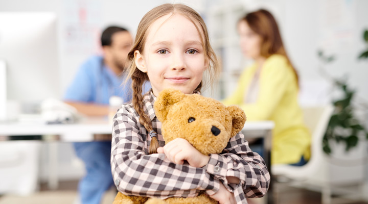 Child holding her teddy bear with parent talking to doctor in background