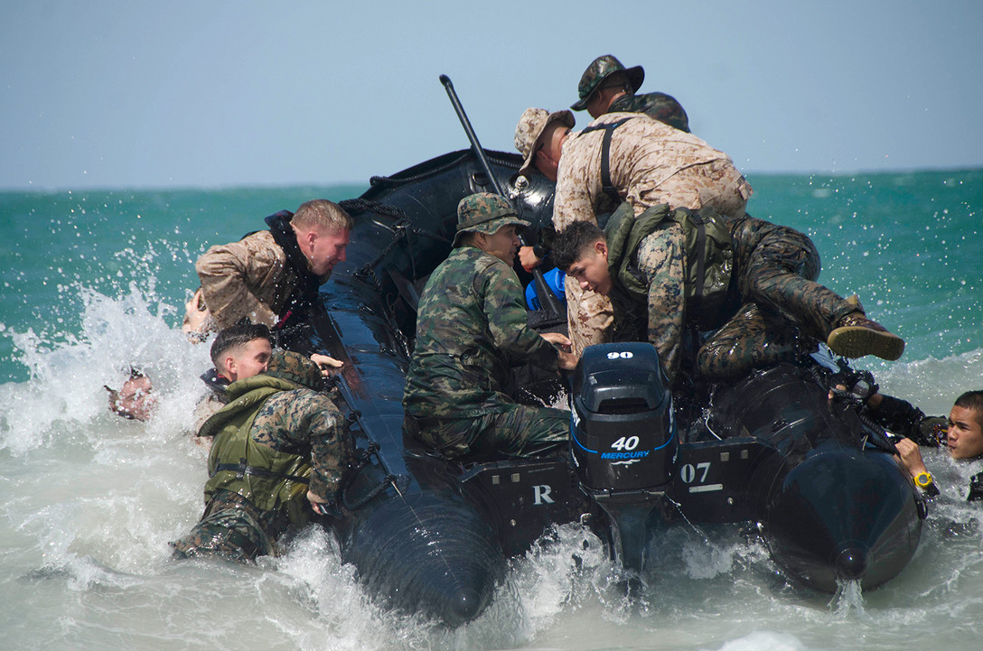 Troops jump onto an inflatable boat maneuvering in white ocean water.