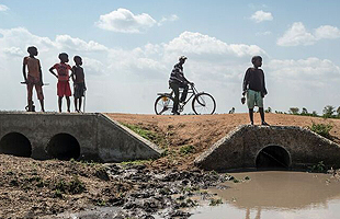 A boy bicycles on a bridge over a field of mud with other children watching