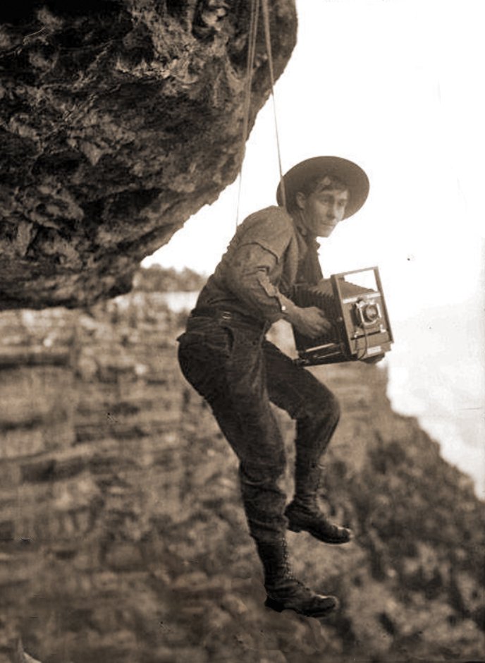 Historic black and white photo of a young man (Pioneer Grand Canyon photographer Emery Kolb) suspended by a rope from a cliff while holding a large view camera. 