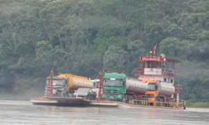 A ferry carrying two oil tankers across a river with lush green tress in the background