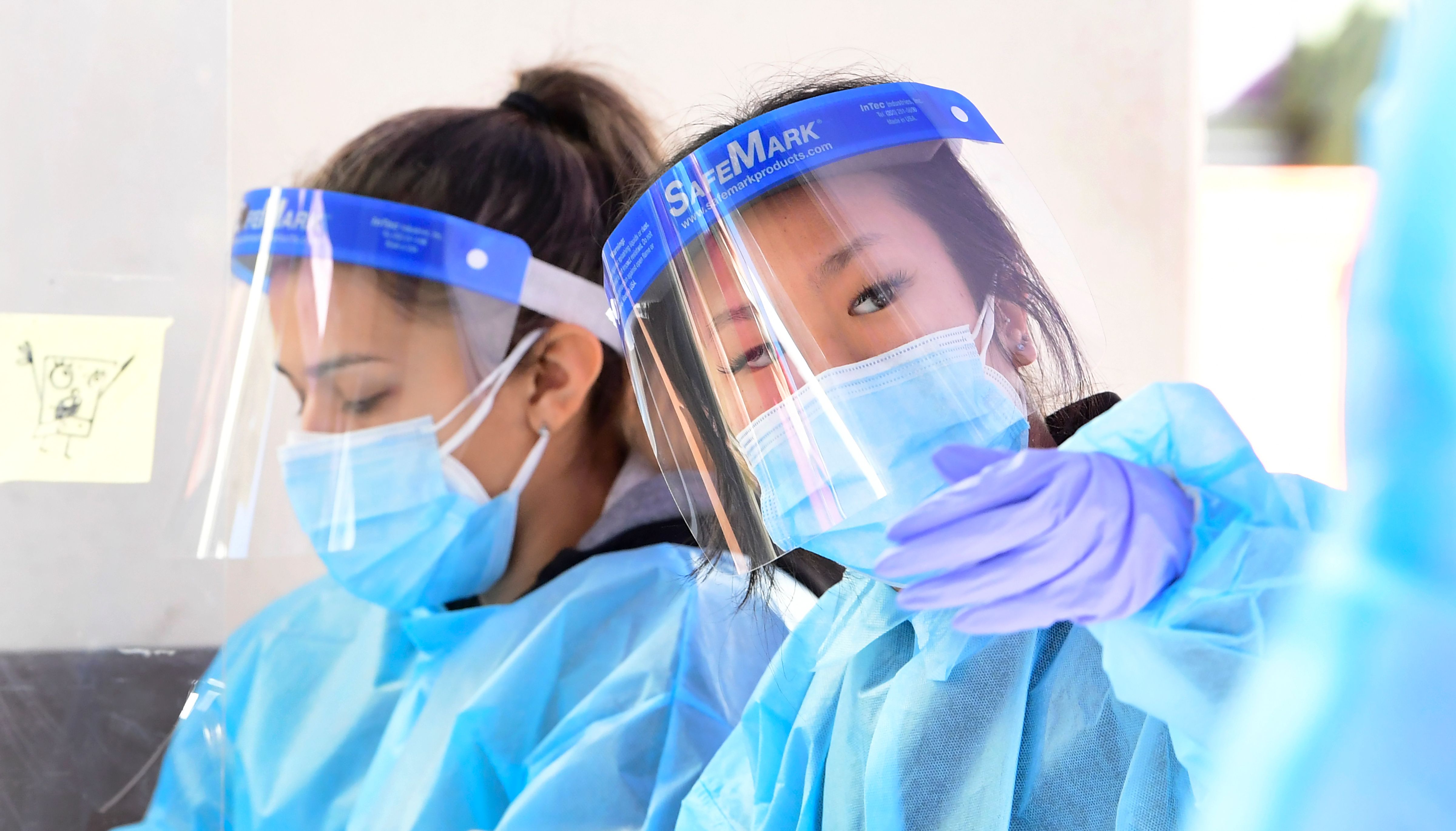 A woman in a blue surgical mask and face shield gestures toward the camera as another woman in the same PPE looks down at forms. Both women have on blue surgical gowns.