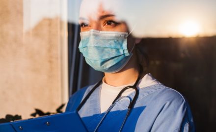 Image for: A woman standing in a window, wearing nurses scrubs and a PPE mask., holding a clip board.