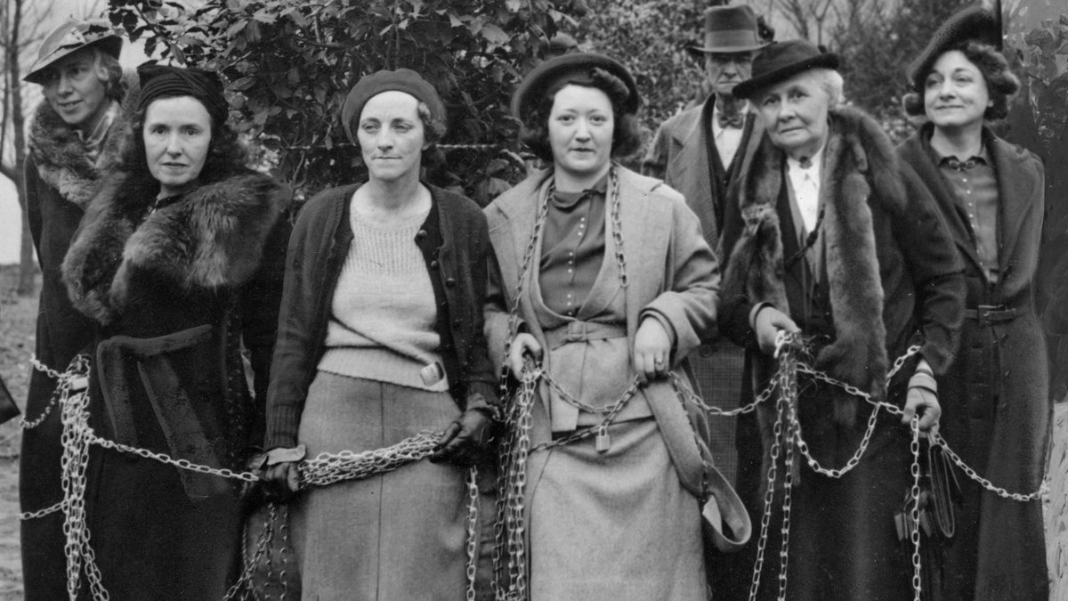 Historic black and white photo of six white women dressed in fancy clothes and hats holding chains and standing in front of a tree.