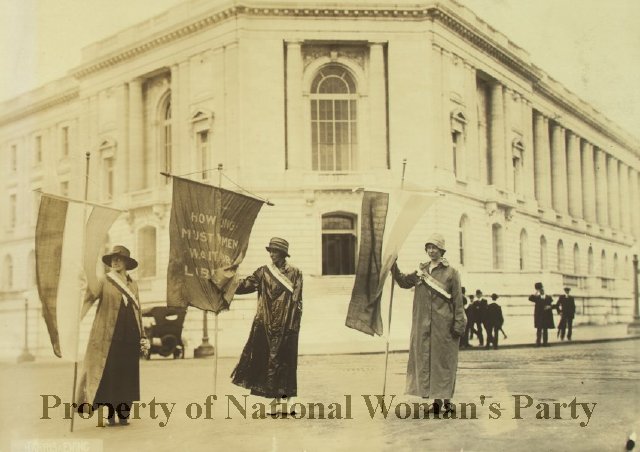 Three women standing outside a building holding banners wearing raincoats, hats, and tricolor suffrage sashes. The middle banner reads "How long must women wait for liberty?" The other two banners are tricolor suffrage banners. Several men look on from a distanc