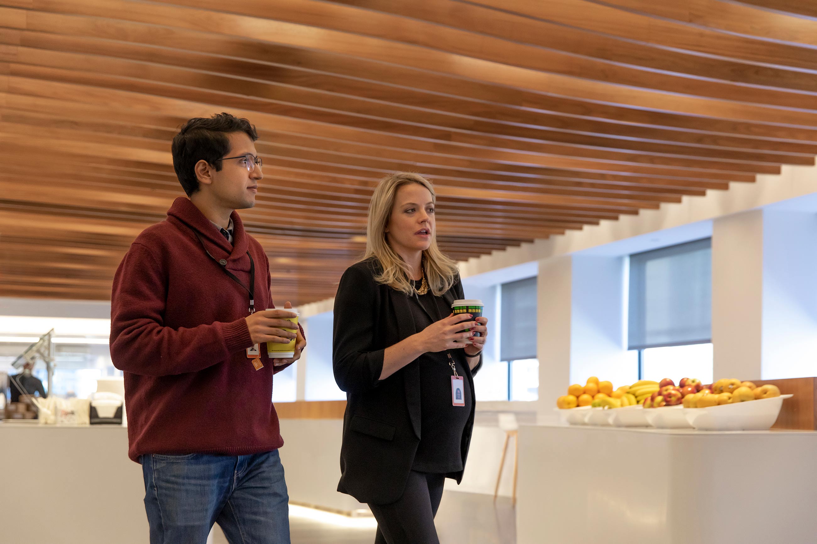 Two Bloomberg employees, one of them pregnant, discuss a project while walking through the office with warm drinks.