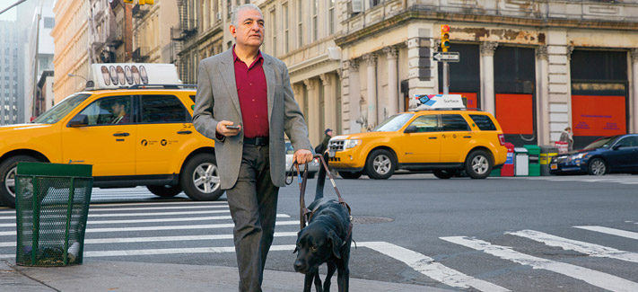 A guide dog leads a well-dressed blind man across a busy city street. The dog appears to be a Labrador.