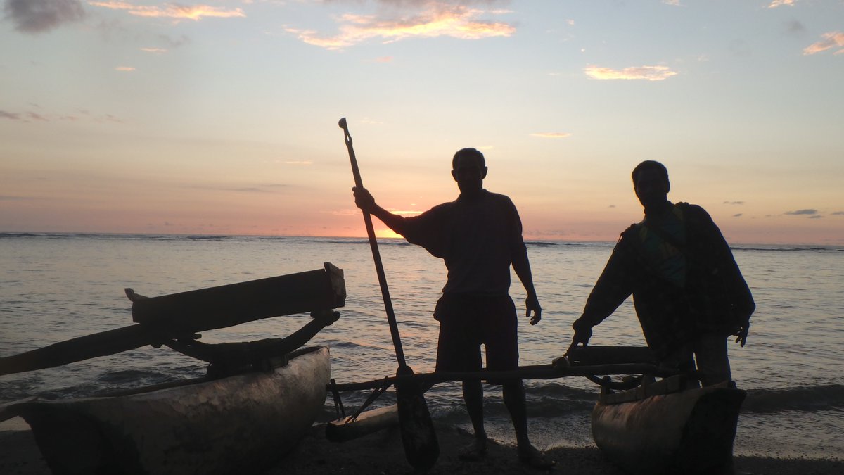 Two men stand by an outrigger canoe, silhouetted in the sunset