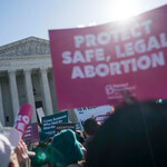 a person in front of the Supreme Court holds a pink Protect Safe, Legal Abortion sing